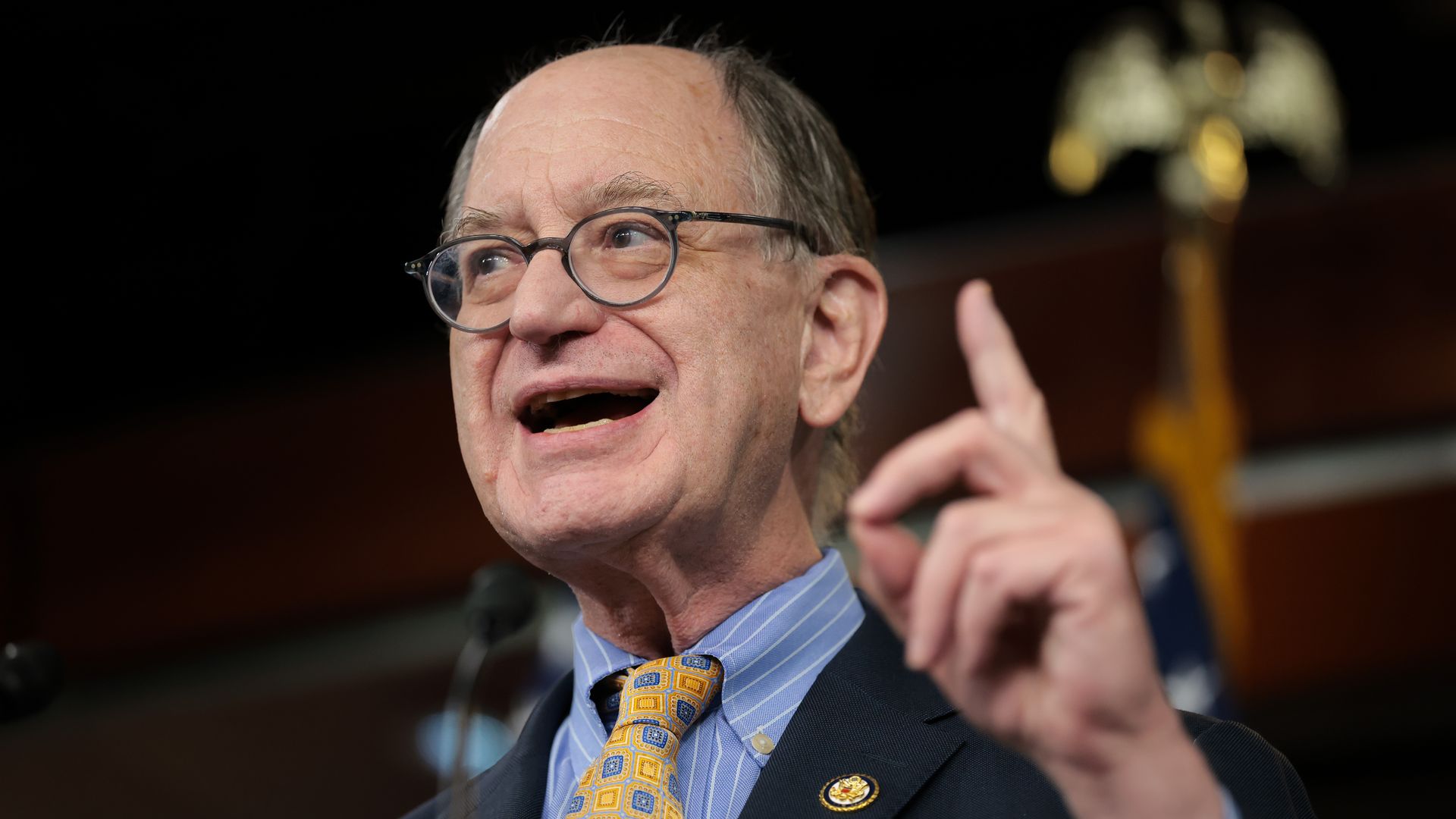 Close-up of a man with round glasses in a dark suit and blue striped shirt, wearing a yellow geometric tie. He is speaking at a podium, with a blurred gold trophy in the background.