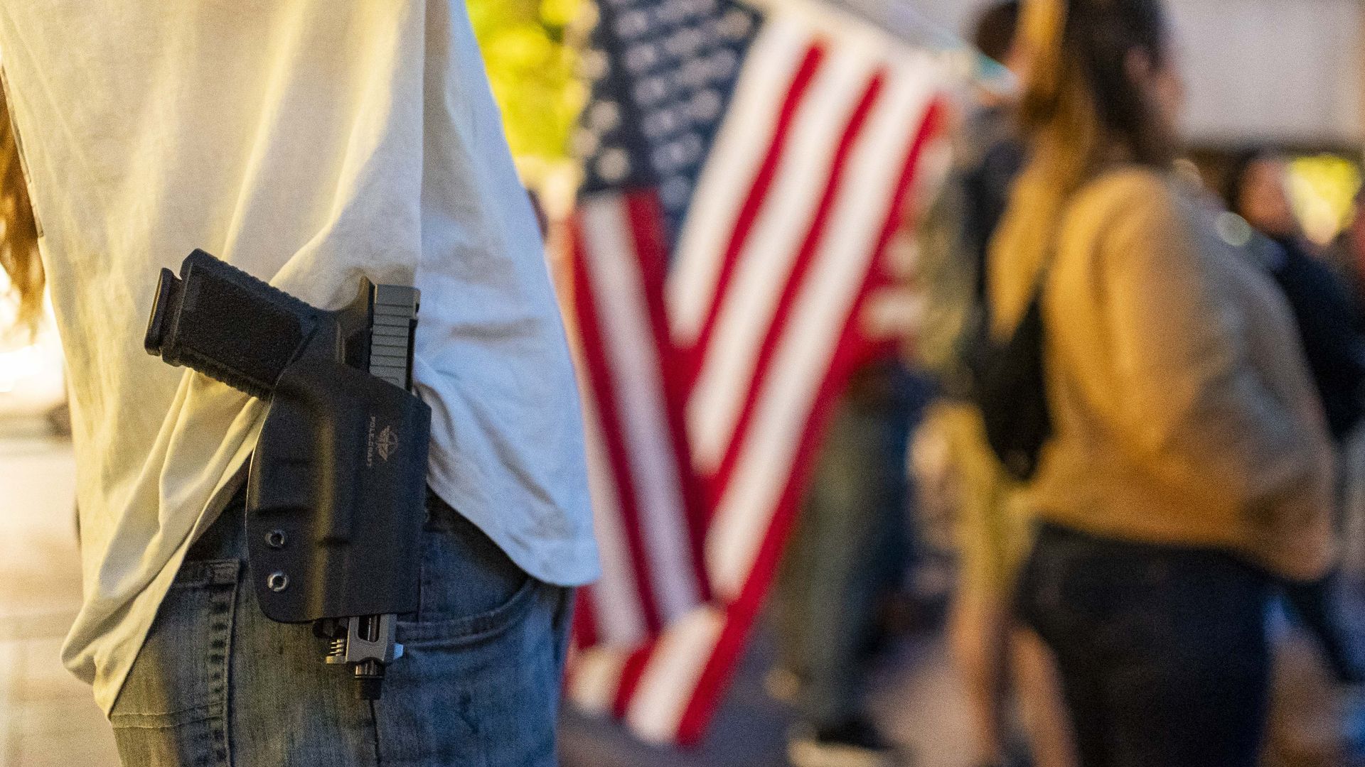 Person wearing jeans and white shirt with a gun on their hip while other people stand behind holding American flags