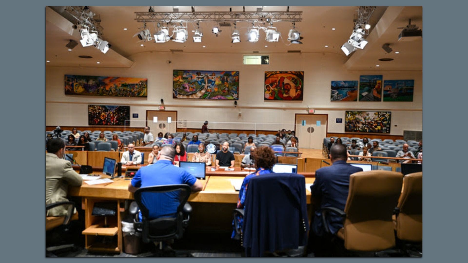 Council chamber or courtroom with a long front desk, people seated at computers; rows of blue chairs filled with attendees; colorful paintings on beige walls and bright overhead lights.