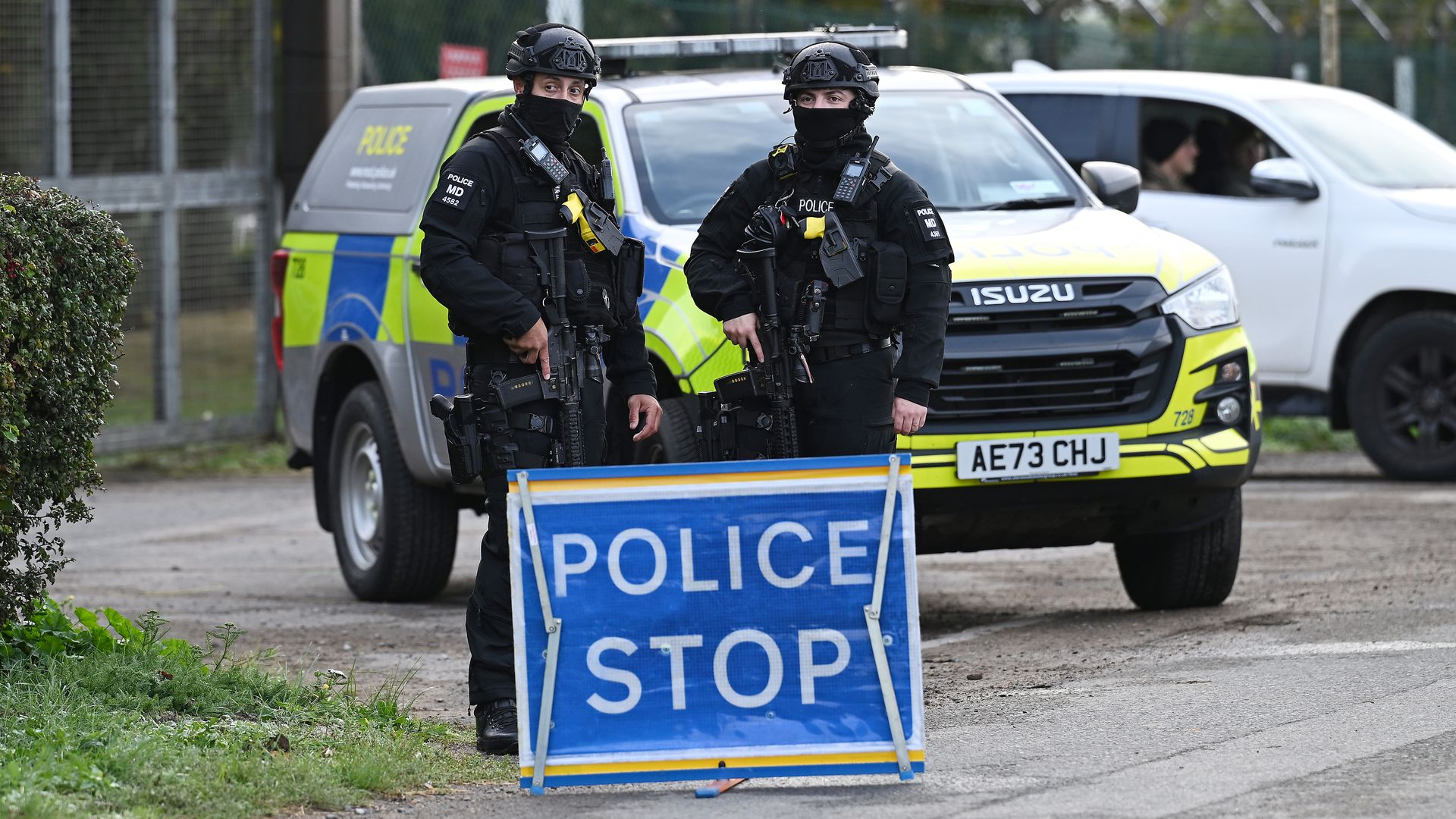 Two armed police officers in black tactical gear and masks stand in front of a yellow and blue police van, a blue police stop sign in the foreground, with a white SUV and fence in the background.