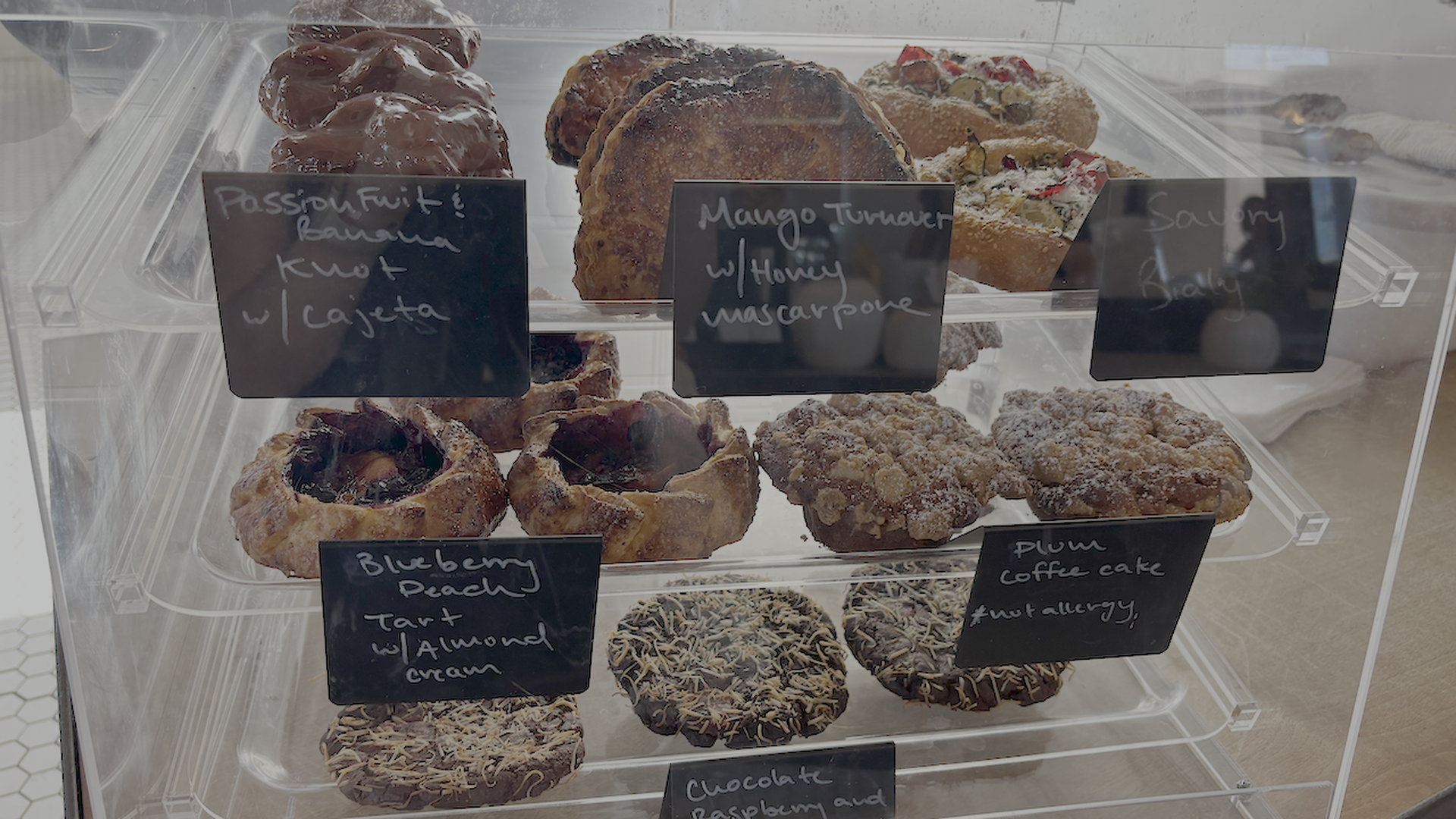Glass bakery display case with donuts and pastries on metal shelves; chalkboard labels read Passion Fruit & Banana, Mango Turnover with Honey Mascarpone, and Blueberry Peach Tart with Almond Cream.