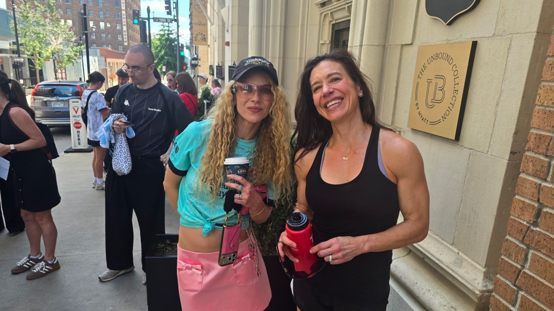 Two smiling women stand on a sidewalk outside Hyatt, The Unbound Collection sign visible. Juno Temple, who plays Keeley in "Ted Lasso," wears turquoise top and pink shorts with a coffee cup; the other, Jenn Higginbotham, in a black tank holds a red bottle.