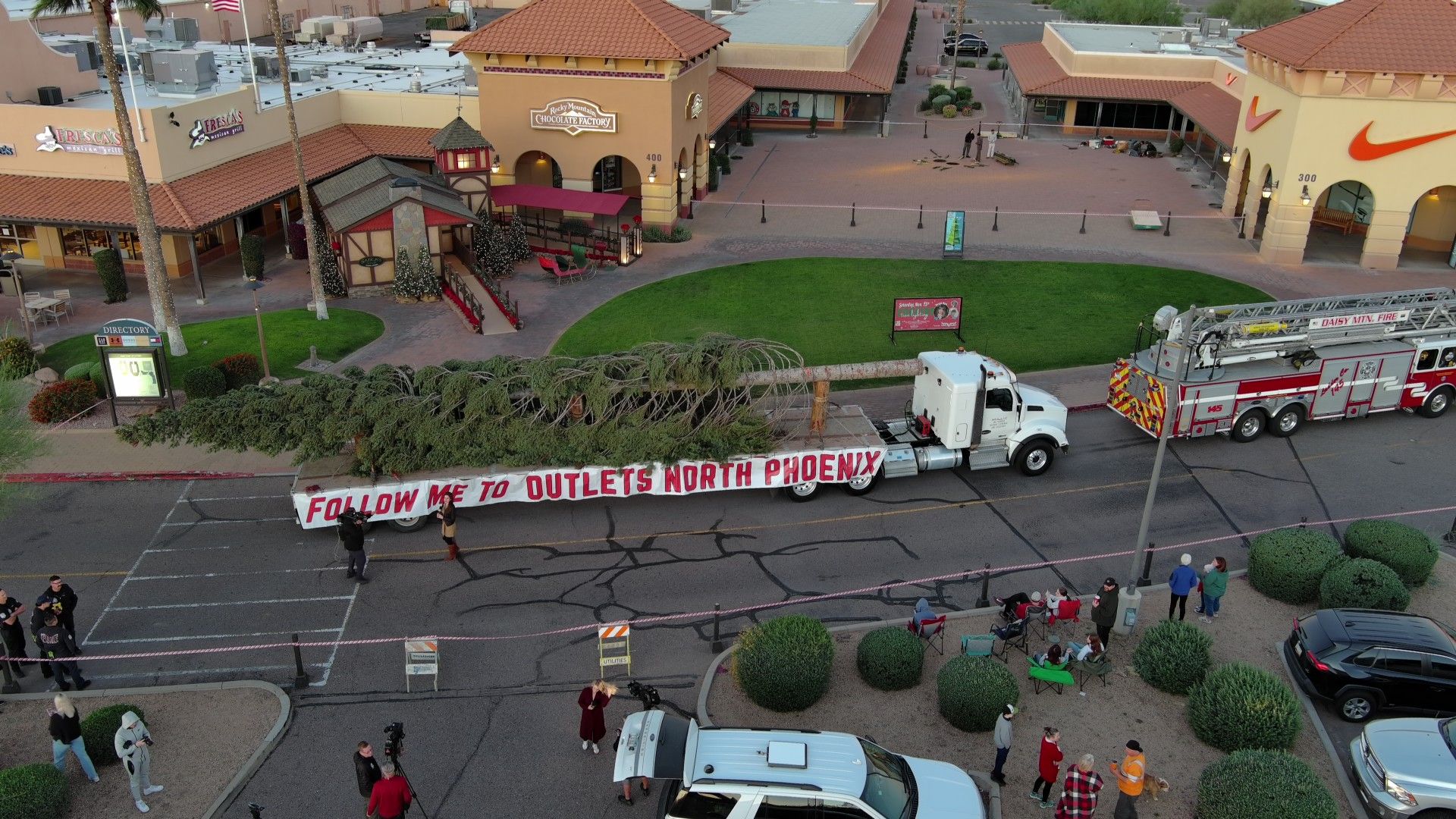 A large Christmas tree on a flatbed truck with a banner reading "FOLLOW ME TO OUTLETS NORTH PHOENIX" is parked in a shopping plaza near stores and a fire truck, with people gathered around.